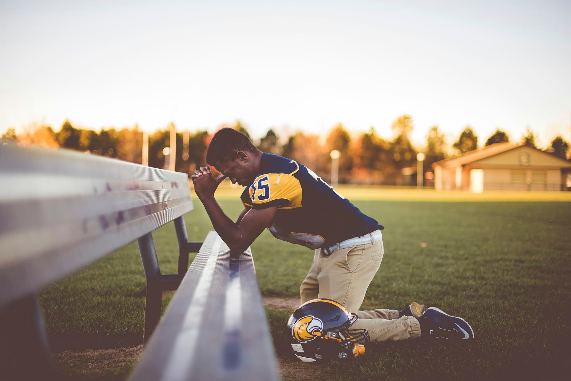 A high school football player kneeling alone at the bench with his head down and his helmet on the grass